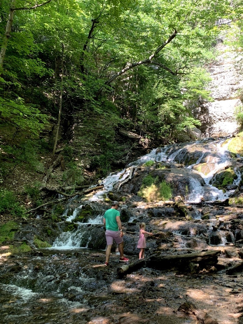 Man in shorts and young girl in pink dress wade in waterfall