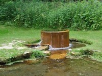 Round iron cistern overflows with water, green foliage behind